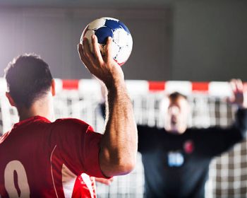 Penalty shoot. Rear view of handball player shooting at goal, while goalkeeper is in the background with his hands raised.